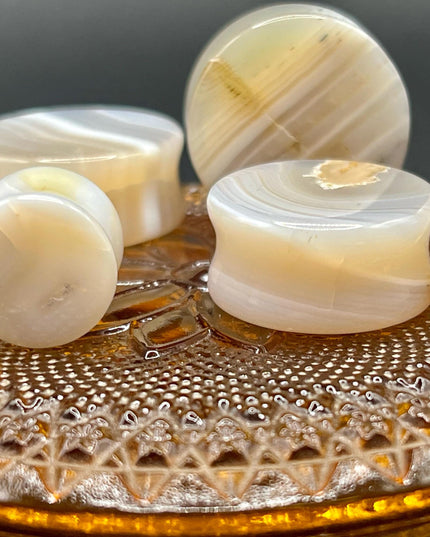 A serene pair of White Line Agate Plugs rests on a soft, neutral background, showcasing their natural, minimalist beauty. Each plug features delicate layers of white, cream, and translucent tones, with subtle linear banding unique to every piece. The polished finish enhances the stone’s soft sheen and calming aesthetic.