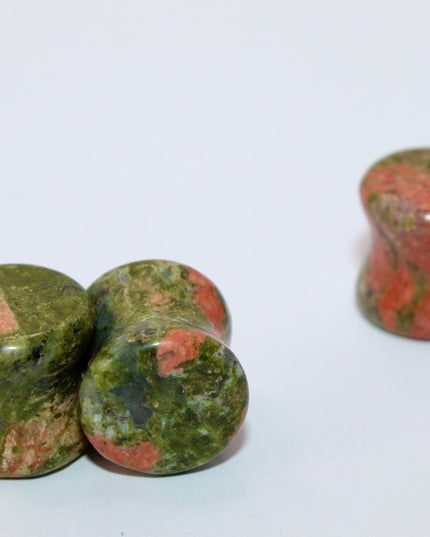 A grounded and vibrant pair of Unakite Double Flare Stone Plugs is displayed on a textured natural background. Each plug showcases the stone’s distinctive blend of mossy green epidote and soft, salmon-pink feldspar—creating a one-of-a-kind marbled pattern that feels both earthy and energizing. The smooth, hand-polished finish enhances the natural contrast in the stone’s texture and color. With a double flare shape for secure, comfortable wear, these plugs combine style with the natural, balancing energy Una