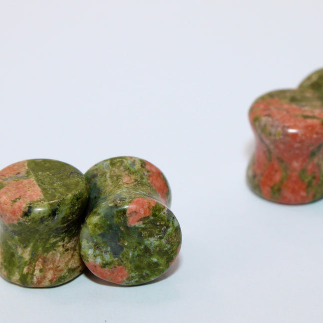 A grounded and vibrant pair of Unakite Double Flare Stone Plugs is displayed on a textured natural background. Each plug showcases the stone’s distinctive blend of mossy green epidote and soft, salmon-pink feldspar—creating a one-of-a-kind marbled pattern that feels both earthy and energizing. The smooth, hand-polished finish enhances the natural contrast in the stone’s texture and color. With a double flare shape for secure, comfortable wear, these plugs combine style with the natural, balancing energy Una