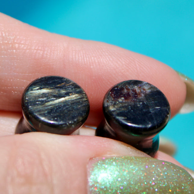 A stunning pair of Astrophyllite FLASH Double Flare Plugs rests against a dark, textured backdrop, their surfaces gleaming with bursts of golden-bronze metallic flash. Each plug is crafted from genuine astrophyllite, a rare and complex stone known for its radiant, starburst-like inclusions that shimmer brilliantly under light. The deep black and charcoal base is layered with reflective veins and flashes of copper, gold, and occasionally blue—giving these plugs a cosmic, otherworldly appearance.