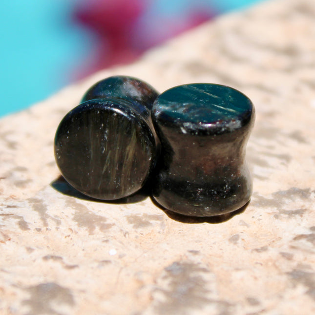 A stunning pair of Astrophyllite FLASH Double Flare Plugs rests against a dark, textured backdrop, their surfaces gleaming with bursts of golden-bronze metallic flash. Each plug is crafted from genuine astrophyllite, a rare and complex stone known for its radiant, starburst-like inclusions that shimmer brilliantly under light. The deep black and charcoal base is layered with reflective veins and flashes of copper, gold, and occasionally blue—giving these plugs a cosmic, otherworldly appearance.