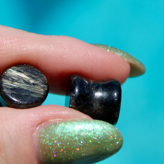 A stunning pair of Astrophyllite FLASH Double Flare Plugs rests against a dark, textured backdrop, their surfaces gleaming with bursts of golden-bronze metallic flash. Each plug is crafted from genuine astrophyllite, a rare and complex stone known for its radiant, starburst-like inclusions that shimmer brilliantly under light. The deep black and charcoal base is layered with reflective veins and flashes of copper, gold, and occasionally blue—giving these plugs a cosmic, otherworldly appearance.