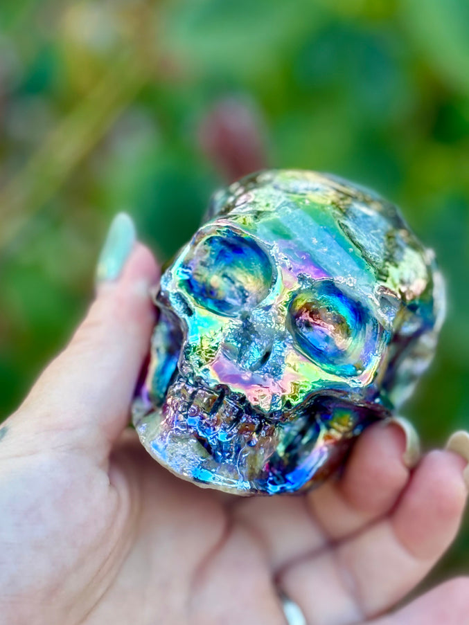 A mesmerizing Genuine Quartz Skull Carving with Rainbow Aura Treatment glows with an ethereal spectrum of colors, resting against a dark, reflective surface. The quartz base is expertly carved into a detailed skull, complete with defined features and a polished finish. The surface shimmers with iridescent hues—vivid blues, purples, pinks, and golds—created through the rainbow aura treatment, a process that bonds precious metals to the quartz for a permanent, luminous effect. 