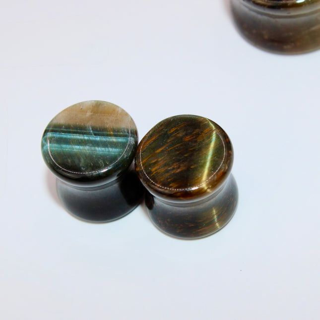 A sleek and polished pair of Blue Tiger Eye plugs, also known as Falcon’s Eye, radiating deep shades of midnight blue, slate, and hints of black. Each plug features the signature chatoyancy of the stone—a silky, light-catching shimmer that moves across the surface as it shifts in the light. The smooth, double-flared design offers both comfort and style, with each plug showcasing natural banding and a unique pattern.