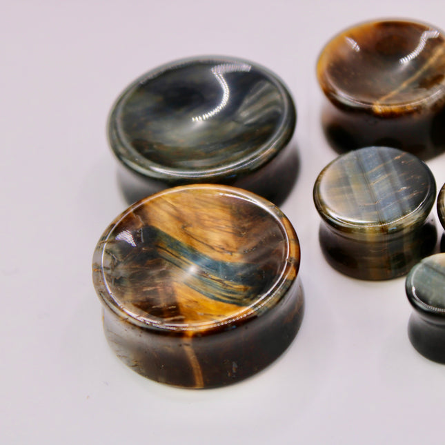 A sleek and polished pair of Blue Tiger Eye plugs, also known as Falcon’s Eye, radiating deep shades of midnight blue, slate, and hints of black. Each plug features the signature chatoyancy of the stone—a silky, light-catching shimmer that moves across the surface as it shifts in the light. The smooth, double-flared design offers both comfort and style, with each plug showcasing natural banding and a unique pattern.