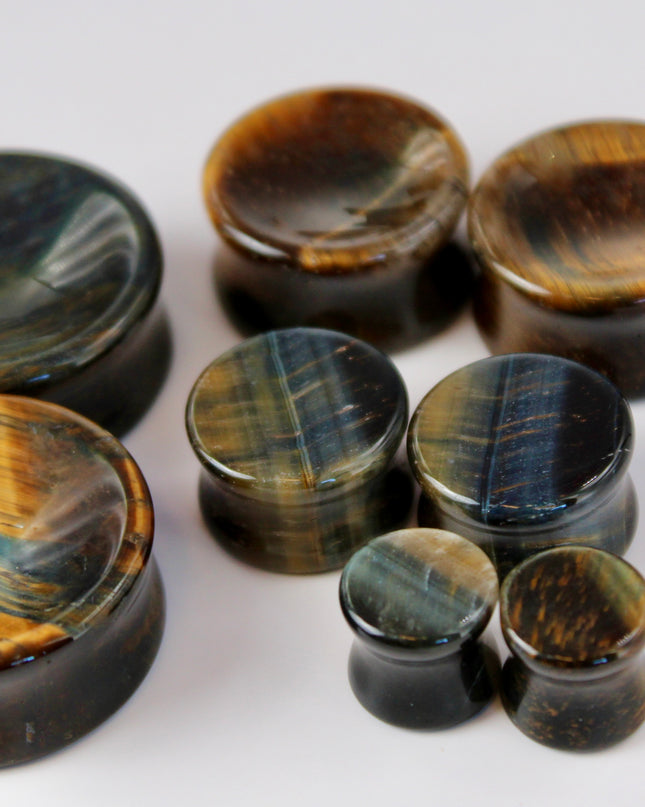 BLUE TIGERS A sleek and polished pair of Blue Tiger Eye plugs, also known as Falcon’s Eye, radiating deep shades of midnight blue, slate, and hints of black. Each plug features the signature chatoyancy of the stone—a silky, light-catching shimmer that moves across the surface as it shifts in the light. The smooth, double-flared design offers both comfort and style, with each plug showcasing natural banding and a unique pattern.