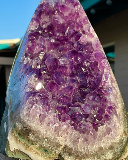 A stunning Amethyst Druzy Cluster Geode Cathedral stands tall with a graceful, arched silhouette reminiscent of gothic architecture. The front face reveals a deep cavern densely lined with sparkling violet and lavender druzy crystals, each point catching the light to create a glittering, celestial effect. The interior displays rich gradients of purple, fading into soft quartz and smoky undertones near the edges. 