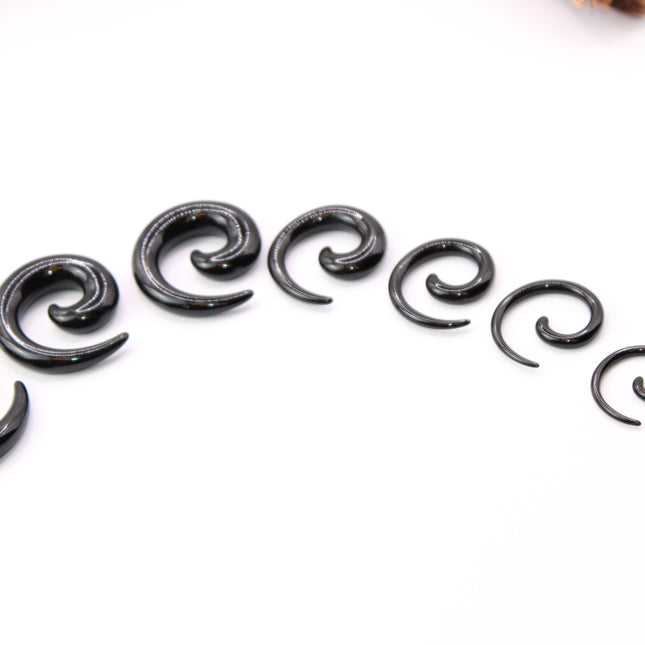 A pair of sleek, black acrylic spiral taper earrings displayed on a clean, neutral surface. The spirals curve smoothly in a bold, symmetrical design, tapering to fine points at the ends. Their glossy finish reflects light subtly, emphasizing their smooth texture and modern, minimalist style. Sized for stretched ears, the earrings showcase a striking balance of simplicity and statement, perfect for alternative or gauged jewelry wearers.