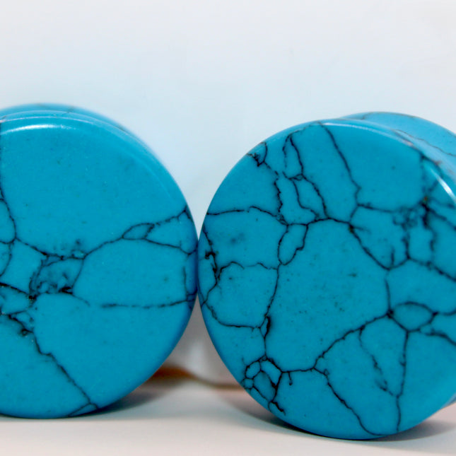 A vibrant pair of Blue Howlite Stone Plugs is shown resting on a soft beige surface. The plugs are round, polished to a high shine, and display a brilliant turquoise-blue color. Each plug is marked with distinctive dark gray to black veining in a web-like pattern—natural to howlite that has been dyed blue to resemble turquoise. The smooth, glossy finish enhances the contrast between the bright blue background and the delicate marbling. The lighting gently reflects off the curved surfaces, emphasizing their