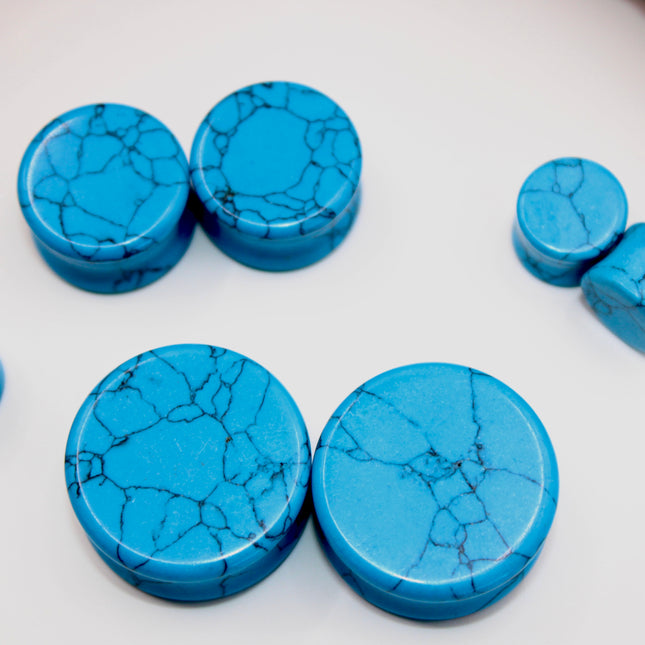 A vibrant pair of Blue Howlite Stone Plugs is shown resting on a soft beige surface. The plugs are round, polished to a high shine, and display a brilliant turquoise-blue color. Each plug is marked with distinctive dark gray to black veining in a web-like pattern—natural to howlite that has been dyed blue to resemble turquoise. The smooth, glossy finish enhances the contrast between the bright blue background and the delicate marbling. The lighting gently reflects off the curved surfaces, emphasizing their 