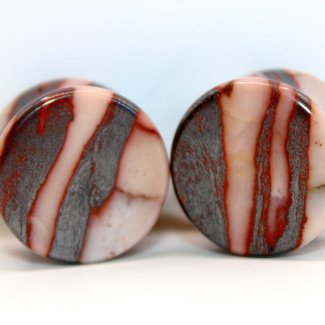 A striking pair of Natural Hematite Brown Double Flared Plugs rests against a soft neutral background, their deep, earthy tones catching the light with a smooth metallic sheen. Each plug is truly one of a kind, showcasing a rich blend of dark brown, bronze, and gunmetal hues with subtle shimmer—hallmarks of natural hematite with unique mineral variations. The double flare design ensures a snug and comfortable fit, while the polished surface adds a sleek, reflective quality. 