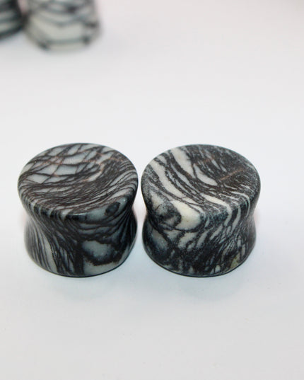 A pair of polished Picasso Stone plugs sits against a neutral-toned background, highlighting their natural beauty. Each plug features a unique marbled pattern of gray, black, tan, and cream tones—resembling abstract brush strokes, much like a miniature piece of modern art. The surface is smooth and glossy, reflecting light subtly. The stone’s intricate veining gives each piece a one-of-a-kind appearance, with soft contrasts and bold lines. The plugs are shaped for comfort and balance, perfect for stretched 