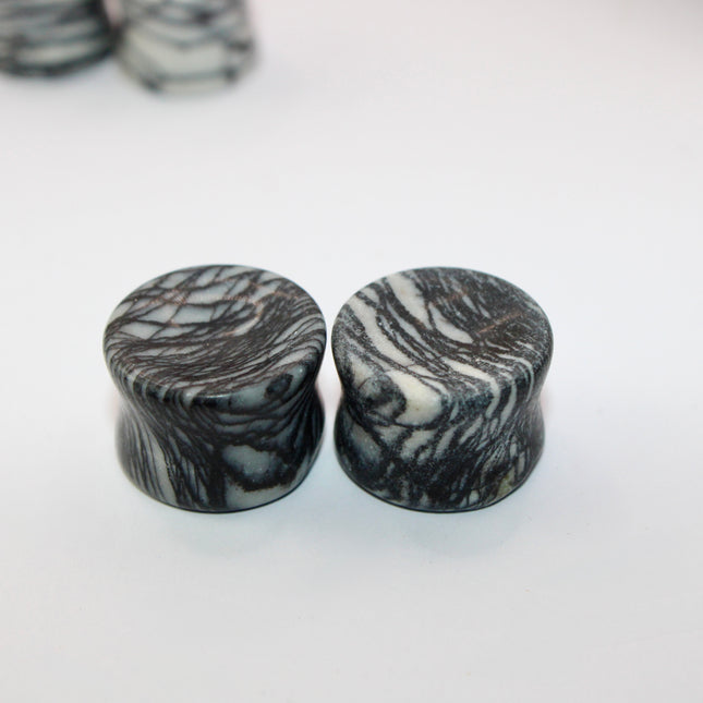 A pair of polished Picasso Stone plugs sits against a neutral-toned background, highlighting their natural beauty. Each plug features a unique marbled pattern of gray, black, tan, and cream tones—resembling abstract brush strokes, much like a miniature piece of modern art. The surface is smooth and glossy, reflecting light subtly. The stone’s intricate veining gives each piece a one-of-a-kind appearance, with soft contrasts and bold lines. The plugs are shaped for comfort and balance, perfect for stretched