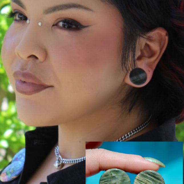 A bold pair of Green Jasper Double Flare Plugs sits elegantly on a neutral surface, radiating natural strength and earthy charm. Each plug is carved from genuine Green Jasper, showcasing deep olive and forest tones, marbled with organic patterns of black, brown, or mossy veining. The polished surface highlights the stone’s natural texture and depth, making every pair unique. 