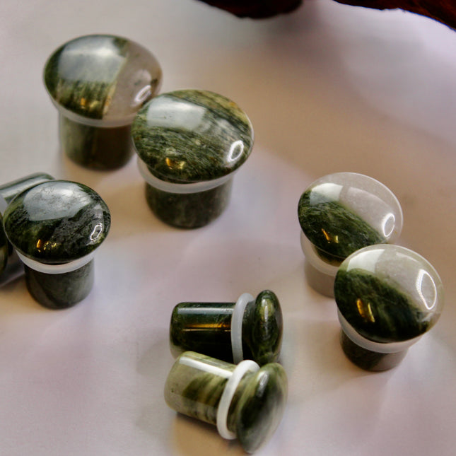 A striking pair of Green Jasper Single Flare Plugs is displayed against an earthy-toned backdrop. Each plug features deep forest green hues with natural patterns of dark veining and subtle swirls, unique to every piece. The surface is smoothly polished, bringing out the rich, organic tones of the stone. Designed with a single flare and a comfortable wearable area, they include O-rings for a secure fit.