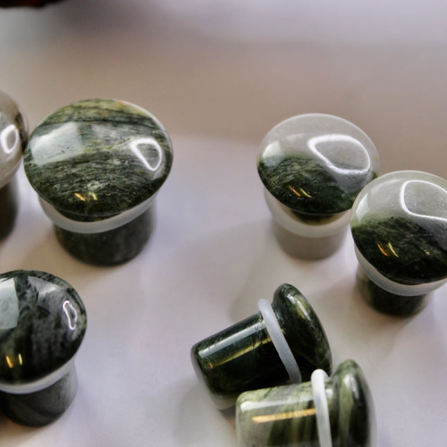 A striking pair of Green Jasper Single Flare Plugs is displayed against an earthy-toned backdrop. Each plug features deep forest green hues with natural patterns of dark veining and subtle swirls, unique to every piece. The surface is smoothly polished, bringing out the rich, organic tones of the stone. Designed with a single flare and a comfortable wearable area, they include O-rings for a secure fit. 