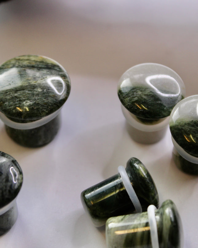 A striking pair of Green Jasper Single Flare Plugs is displayed against an earthy-toned backdrop. Each plug features deep forest green hues with natural patterns of dark veining and subtle swirls, unique to every piece. The surface is smoothly polished, bringing out the rich, organic tones of the stone. Designed with a single flare and a comfortable wearable area, they include O-rings for a secure fit. 