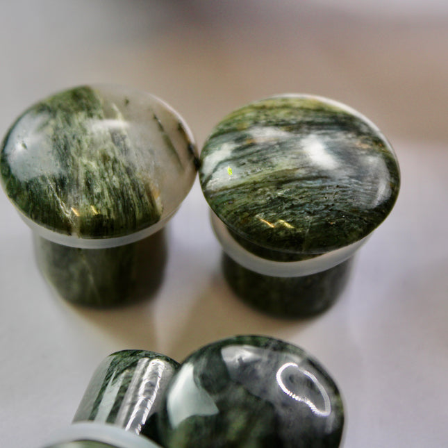 A striking pair of Green Jasper Single Flare Plugs is displayed against an earthy-toned backdrop. Each plug features deep forest green hues with natural patterns of dark veining and subtle swirls, unique to every piece. The surface is smoothly polished, bringing out the rich, organic tones of the stone. Designed with a single flare and a comfortable wearable area, they include O-rings for a secure fit. 