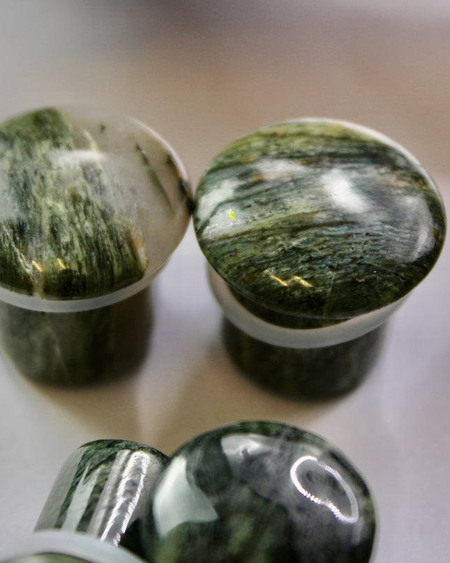 A striking pair of Green Jasper Single Flare Plugs is displayed against an earthy-toned backdrop. Each plug features deep forest green hues with natural patterns of dark veining and subtle swirls, unique to every piece. The surface is smoothly polished, bringing out the rich, organic tones of the stone. Designed with a single flare and a comfortable wearable area, they include O-rings for a secure fit. 