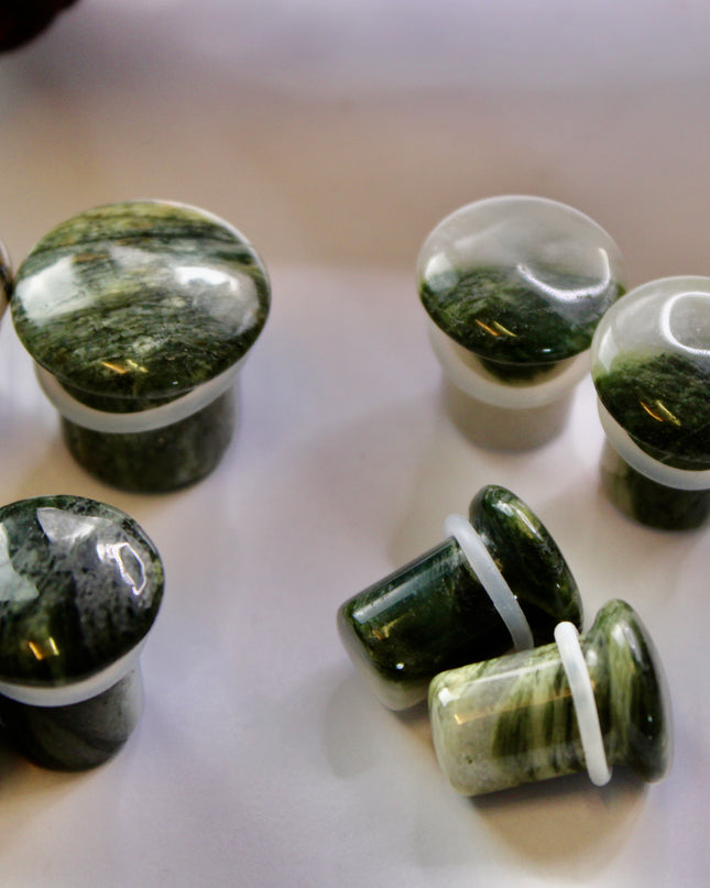 A striking pair of Green Jasper Single Flare Plugs is displayed against an earthy-toned backdrop. Each plug features deep forest green hues with natural patterns of dark veining and subtle swirls, unique to every piece. The surface is smoothly polished, bringing out the rich, organic tones of the stone. Designed with a single flare and a comfortable wearable area, they include O-rings for a secure fit. 