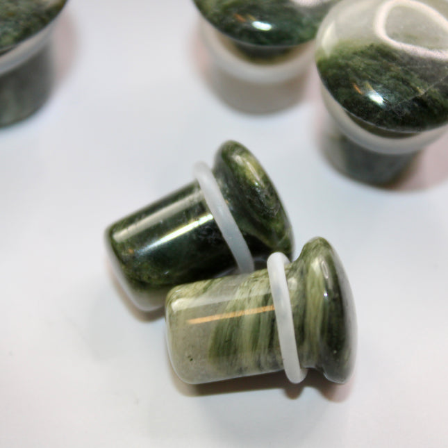 A striking pair of Green Jasper Single Flare Plugs is displayed against an earthy-toned backdrop. Each plug features deep forest green hues with natural patterns of dark veining and subtle swirls, unique to every piece. The surface is smoothly polished, bringing out the rich, organic tones of the stone. Designed with a single flare and a comfortable wearable area, they include O-rings for a secure fit.