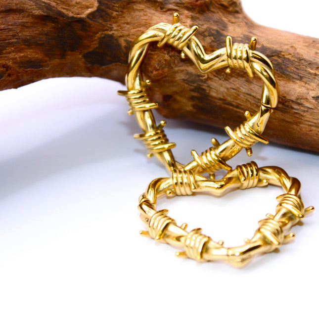 A striking pair of heart-shaped hangers designed for stretched ears, crafted in a shiny gold-tone metal with detailed barbed wire accents wrapping around the heart outline. The edgy design blends romance with rebellion, featuring smooth curves contrasted by sharp, sculpted wire barbs for a bold, punk-inspired aesthetic. Made to hang comfortably in stretched lobes, these hangers are lightweight yet visually impactful—perfect for expressing individuality with attitude and style.