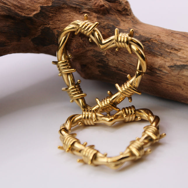 A striking pair of heart-shaped hangers designed for stretched ears, crafted in a shiny gold-tone metal with detailed barbed wire accents wrapping around the heart outline. The edgy design blends romance with rebellion, featuring smooth curves contrasted by sharp, sculpted wire barbs for a bold, punk-inspired aesthetic. Made to hang comfortably in stretched lobes, these hangers are lightweight yet visually impactful—perfect for expressing individuality with attitude and style.
