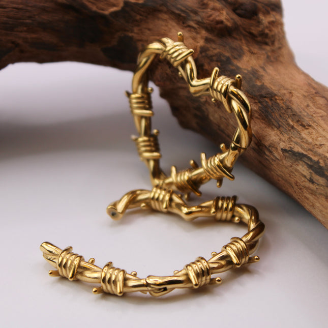 A striking pair of heart-shaped hangers designed for stretched ears, crafted in a shiny gold-tone metal with detailed barbed wire accents wrapping around the heart outline. The edgy design blends romance with rebellion, featuring smooth curves contrasted by sharp, sculpted wire barbs for a bold, punk-inspired aesthetic. Made to hang comfortably in stretched lobes, these hangers are lightweight yet visually impactful—perfect for expressing individuality with attitude and style.