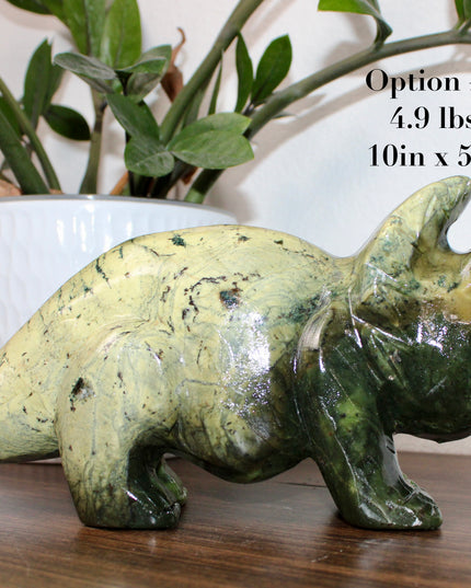 Green stone triceratops figurine on a wooden surface with plants in the background