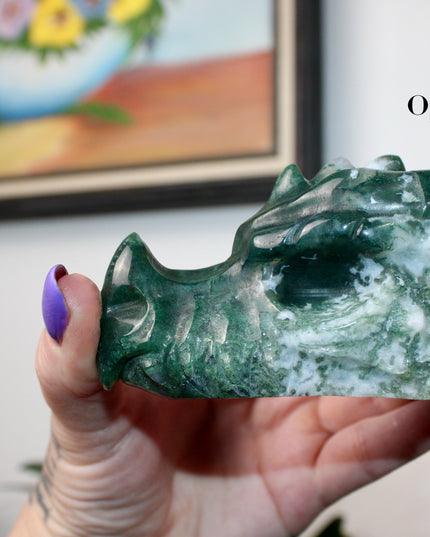 Bring a sense of majestic artistry and natural wonder into your space with this hand‑carved moss agate dragon skull. Sculpted from a single piece of genuine moss agate celebrated for its swirling green “moss-like” inclusions set against milky chalcedony, each piece showcases its own unique, earthy landscape