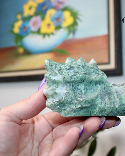 Bring a sense of majestic artistry and natural wonder into your space with this hand‑carved moss agate dragon skull. Sculpted from a single piece of genuine moss agate celebrated for its swirling green “moss-like” inclusions set against milky chalcedony, each piece showcases its own unique, earthy landscape