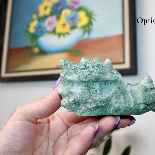 Bring a sense of majestic artistry and natural wonder into your space with this hand‑carved moss agate dragon skull. Sculpted from a single piece of genuine moss agate celebrated for its swirling green “moss-like” inclusions set against milky chalcedony, each piece showcases its own unique, earthy landscape