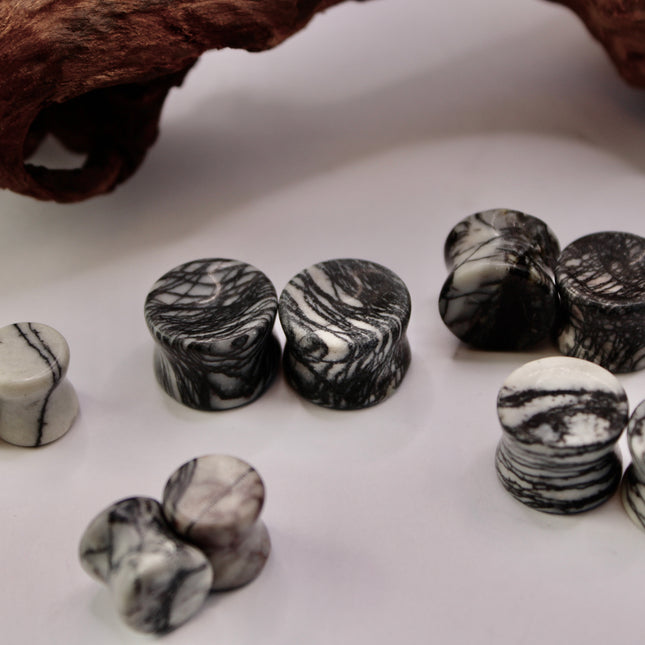 A pair of polished Picasso Stone plugs sits against a neutral-toned background, highlighting their natural beauty. Each plug features a unique marbled pattern of gray, black, tan, and cream tones—resembling abstract brush strokes, much like a miniature piece of modern art. The surface is smooth and glossy, reflecting light subtly. The stone’s intricate veining gives each piece a one-of-a-kind appearance, with soft contrasts and bold lines. The plugs are shaped for comfort and balance, perfect for stretched 