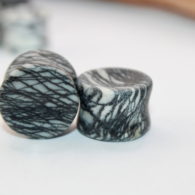 A pair of polished Picasso Stone plugs sits against a neutral-toned background, highlighting their natural beauty. Each plug features a unique marbled pattern of gray, black, tan, and cream tones—resembling abstract brush strokes, much like a miniature piece of modern art. The surface is smooth and glossy, reflecting light subtly. The stone’s intricate veining gives each piece a one-of-a-kind appearance, with soft contrasts and bold lines. The plugs are shaped for comfort and balance, perfect for stretched