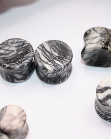 A pair of polished Picasso Stone plugs sits against a neutral-toned background, highlighting their natural beauty. Each plug features a unique marbled pattern of gray, black, tan, and cream tones—resembling abstract brush strokes, much like a miniature piece of modern art. The surface is smooth and glossy, reflecting light subtly. The stone’s intricate veining gives each piece a one-of-a-kind appearance, with soft contrasts and bold lines. The plugs are shaped for comfort and balance, perfect for stretched 
