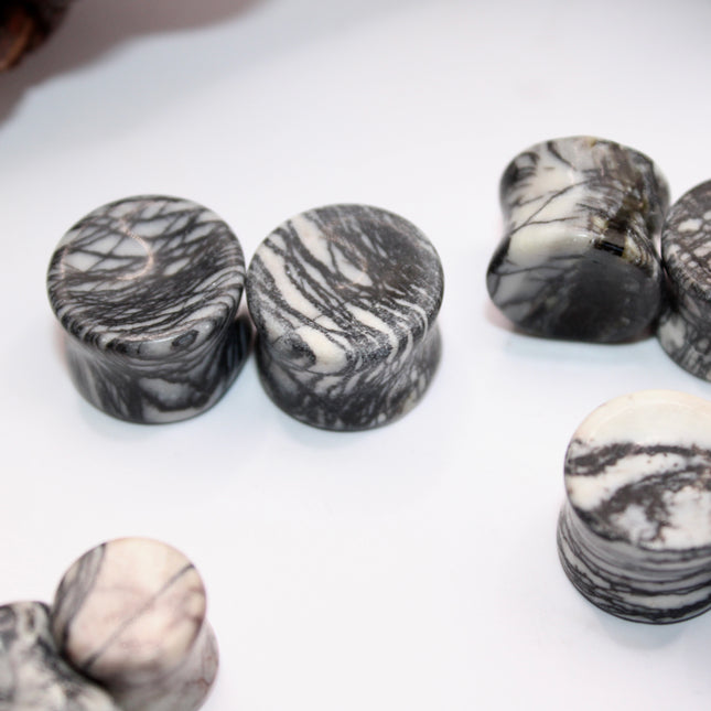 A pair of polished Picasso Stone plugs sits against a neutral-toned background, highlighting their natural beauty. Each plug features a unique marbled pattern of gray, black, tan, and cream tones—resembling abstract brush strokes, much like a miniature piece of modern art. The surface is smooth and glossy, reflecting light subtly. The stone’s intricate veining gives each piece a one-of-a-kind appearance, with soft contrasts and bold lines. The plugs are shaped for comfort and balance, perfect for stretched