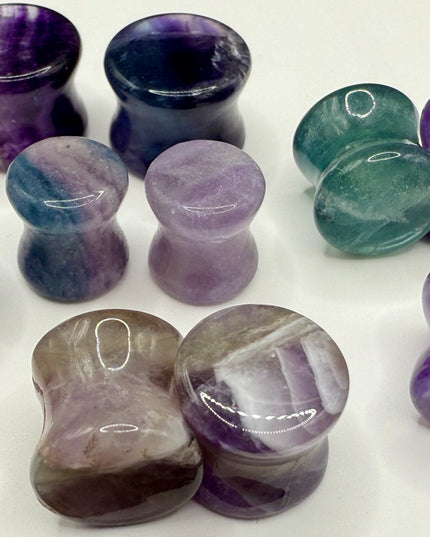 A stunning pair of Rainbow Fluorite stone plugs, expertly carved and polished to a smooth, glossy finish. Each plug displays the natural beauty of fluorite with vivid bands of purple, green, and teal, creating a mesmerizing, layered effect unique to each piece. The translucent crystal catches and reflects light, enhancing its vibrant colors and depth. Designed with a comfortable double flare for a secure fit, these natural crystal gauges are perfect for stretched ears