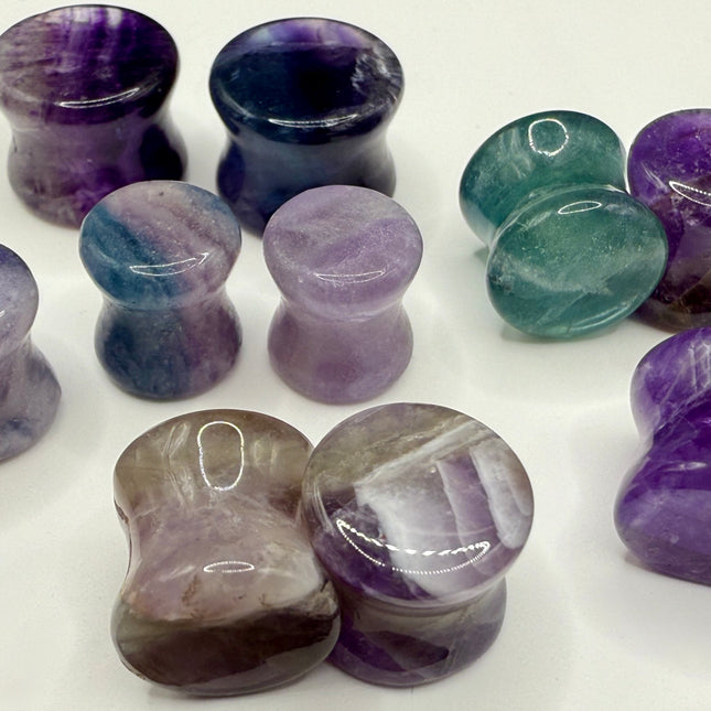 A stunning pair of Rainbow Fluorite stone plugs, expertly carved and polished to a smooth, glossy finish. Each plug displays the natural beauty of fluorite with vivid bands of purple, green, and teal, creating a mesmerizing, layered effect unique to each piece. The translucent crystal catches and reflects light, enhancing its vibrant colors and depth. Designed with a comfortable double flare for a secure fit, these natural crystal gauges are perfect for stretched ears