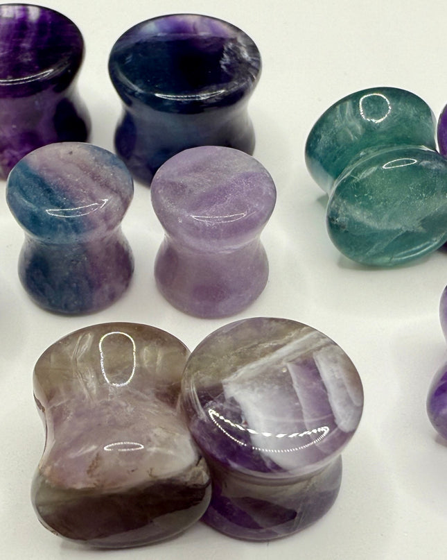 A stunning pair of Rainbow Fluorite stone plugs, expertly carved and polished to a smooth, glossy finish. Each plug displays the natural beauty of fluorite with vivid bands of purple, green, and teal, creating a mesmerizing, layered effect unique to each piece. The translucent crystal catches and reflects light, enhancing its vibrant colors and depth. Designed with a comfortable double flare for a secure fit, these natural crystal gauges are perfect for stretched ears