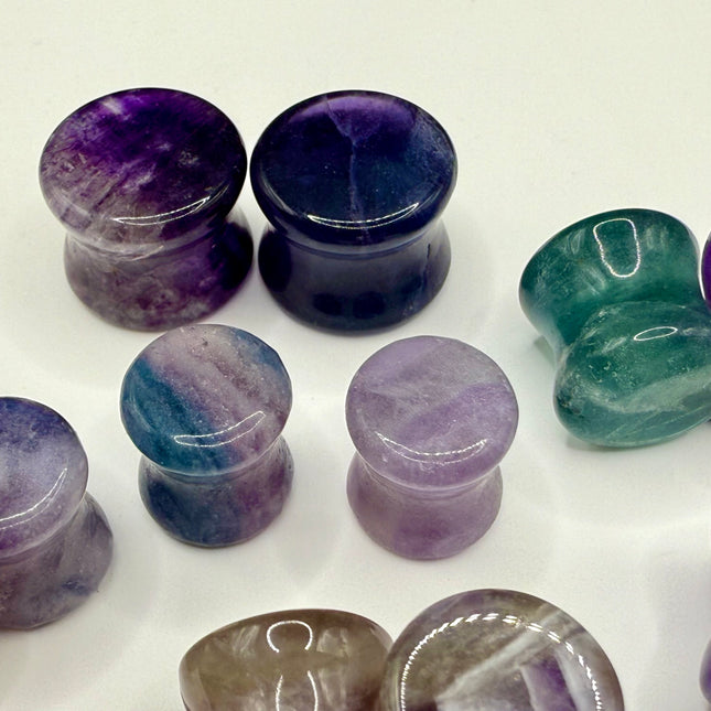 A stunning pair of Rainbow Fluorite stone plugs, expertly carved and polished to a smooth, glossy finish. Each plug displays the natural beauty of fluorite with vivid bands of purple, green, and teal, creating a mesmerizing, layered effect unique to each piece. The translucent crystal catches and reflects light, enhancing its vibrant colors and depth. Designed with a comfortable double flare for a secure fit, these natural crystal gauges are perfect for stretched ears