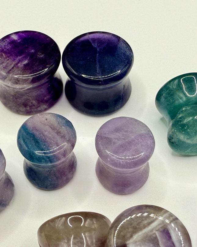 A stunning pair of Rainbow Fluorite stone plugs, expertly carved and polished to a smooth, glossy finish. Each plug displays the natural beauty of fluorite with vivid bands of purple, green, and teal, creating a mesmerizing, layered effect unique to each piece. The translucent crystal catches and reflects light, enhancing its vibrant colors and depth. Designed with a comfortable double flare for a secure fit, these natural crystal gauges are perfect for stretched ears