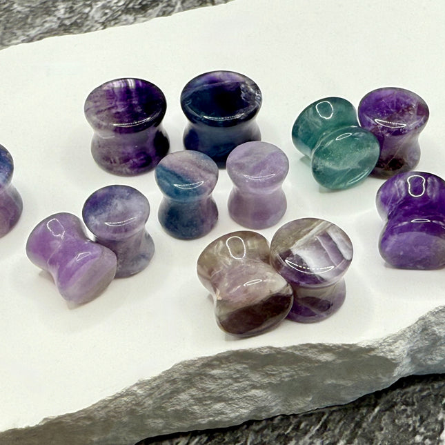 A stunning pair of Rainbow Fluorite stone plugs, expertly carved and polished to a smooth, glossy finish. Each plug displays the natural beauty of fluorite with vivid bands of purple, green, and teal, creating a mesmerizing, layered effect unique to each piece. The translucent crystal catches and reflects light, enhancing its vibrant colors and depth. Designed with a comfortable double flare for a secure fit, these natural crystal gauges are perfect for stretched ears