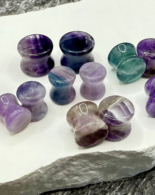 A stunning pair of Rainbow Fluorite stone plugs, expertly carved and polished to a smooth, glossy finish. Each plug displays the natural beauty of fluorite with vivid bands of purple, green, and teal, creating a mesmerizing, layered effect unique to each piece. The translucent crystal catches and reflects light, enhancing its vibrant colors and depth. Designed with a comfortable double flare for a secure fit, these natural crystal gauges are perfect for stretched ears