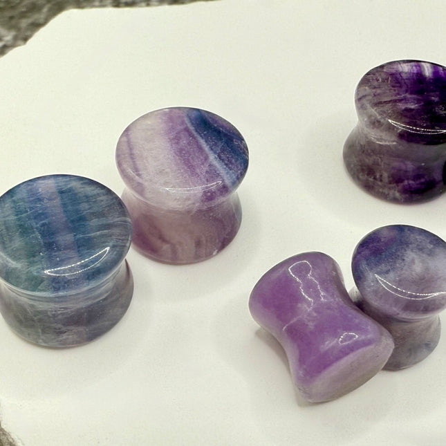 A stunning pair of Rainbow Fluorite stone plugs, expertly carved and polished to a smooth, glossy finish. Each plug displays the natural beauty of fluorite with vivid bands of purple, green, and teal, creating a mesmerizing, layered effect unique to each piece. The translucent crystal catches and reflects light, enhancing its vibrant colors and depth. Designed with a comfortable double flare for a secure fit, these natural crystal gauges are perfect for stretched ears