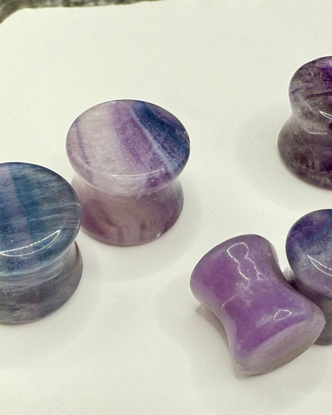 A stunning pair of Rainbow Fluorite stone plugs, expertly carved and polished to a smooth, glossy finish. Each plug displays the natural beauty of fluorite with vivid bands of purple, green, and teal, creating a mesmerizing, layered effect unique to each piece. The translucent crystal catches and reflects light, enhancing its vibrant colors and depth. Designed with a comfortable double flare for a secure fit, these natural crystal gauges are perfect for stretched ears