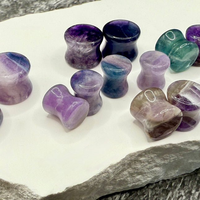 A stunning pair of Rainbow Fluorite stone plugs, expertly carved and polished to a smooth, glossy finish. Each plug displays the natural beauty of fluorite with vivid bands of purple, green, and teal, creating a mesmerizing, layered effect unique to each piece. The translucent crystal catches and reflects light, enhancing its vibrant colors and depth. Designed with a comfortable double flare for a secure fit, these natural crystal gauges are perfect for stretched ears