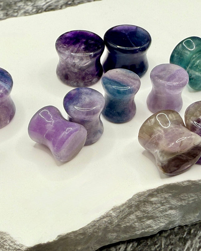 A stunning pair of Rainbow Fluorite stone plugs, expertly carved and polished to a smooth, glossy finish. Each plug displays the natural beauty of fluorite with vivid bands of purple, green, and teal, creating a mesmerizing, layered effect unique to each piece. The translucent crystal catches and reflects light, enhancing its vibrant colors and depth. Designed with a comfortable double flare for a secure fit, these natural crystal gauges are perfect for stretched ears