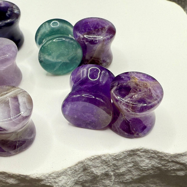 A stunning pair of Rainbow Fluorite stone plugs, expertly carved and polished to a smooth, glossy finish. Each plug displays the natural beauty of fluorite with vivid bands of purple, green, and teal, creating a mesmerizing, layered effect unique to each piece. The translucent crystal catches and reflects light, enhancing its vibrant colors and depth. Designed with a comfortable double flare for a secure fit, these natural crystal gauges are perfect for stretched ears