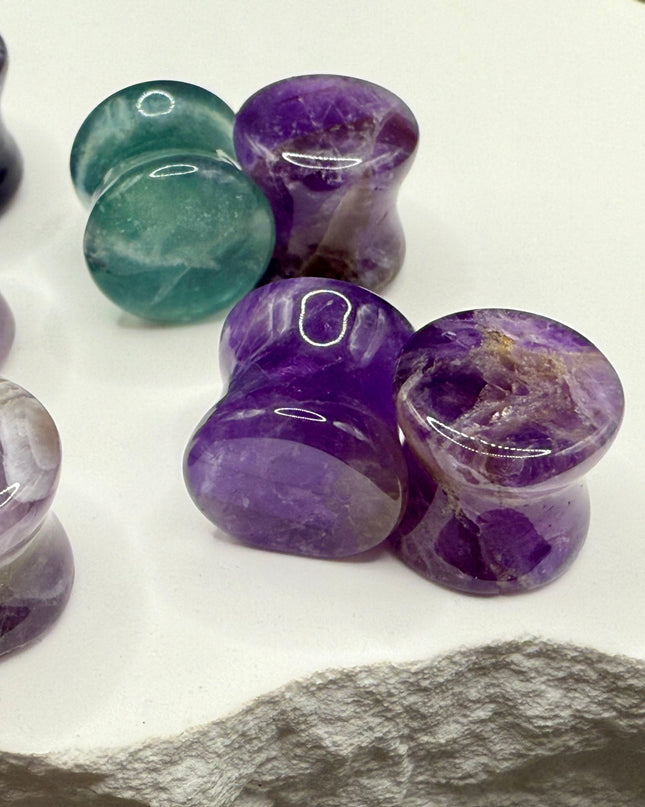 A stunning pair of Rainbow Fluorite stone plugs, expertly carved and polished to a smooth, glossy finish. Each plug displays the natural beauty of fluorite with vivid bands of purple, green, and teal, creating a mesmerizing, layered effect unique to each piece. The translucent crystal catches and reflects light, enhancing its vibrant colors and depth. Designed with a comfortable double flare for a secure fit, these natural crystal gauges are perfect for stretched ears
