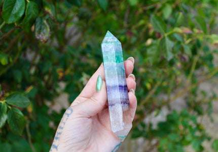 A mesmerizing Rainbow Fluorite Double Terminated Wand lies on a clean, light surface, glowing with layers of vivid color. The wand features natural bands of purple, green, teal, and clear fluorite, stacked like waves of translucent energy. Both ends are precisely cut to pointed terminations, ideal for directing energy in crystal healing or meditation practices. The polished finish enhances the crystal’s clarity and color shifts, with each piece showcasing its own unique pattern. 